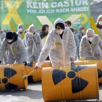 Aktivisten rollen Fässer mit Atomkraft-zeichen auf dem Boden, Sie tragen Schutzkleidung und Brillen. Im Hintergrund ein grünes Banner mit der Aufschrift" Kein Castor von Jülich nach Ahaus!"