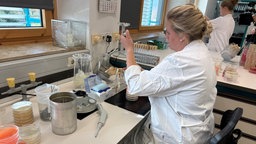 Ein Frau untersucht in einem eißen Kittel etwas in einem Labor. um Sie herum Pipette, petrischalen und Reagenzgläser