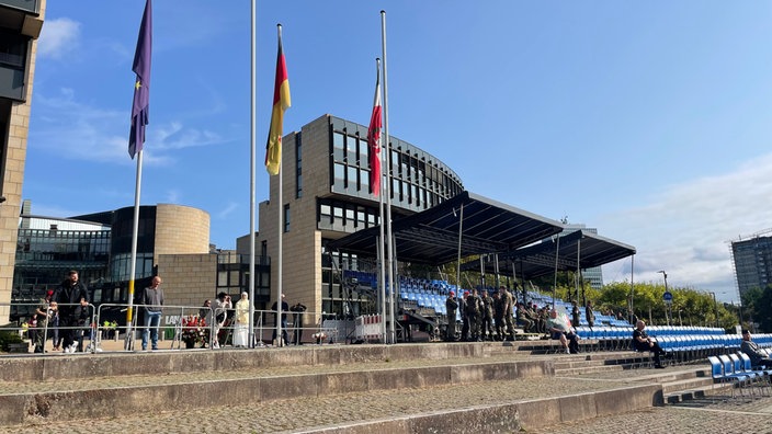 Tribüne mit blauen Sitzen aufgebaut vor dem NRW Landtag. Rekruten und Zuschauer stehen herum. Die Flaggen vor dem NRW Landtag und im Hintergrund das Gebäude.