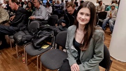 Paula Graf (16) ist bei den Grünen und sitzt in einem Veranstaltungssaal auf einem Stuhl im Publikum