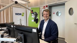 Henning Höne sitzt an einem Tisch mit Mikrofon in einem Radiostudio, vor ihm zwei Bildschirme, im Hintergrund das WDR5-Logo.