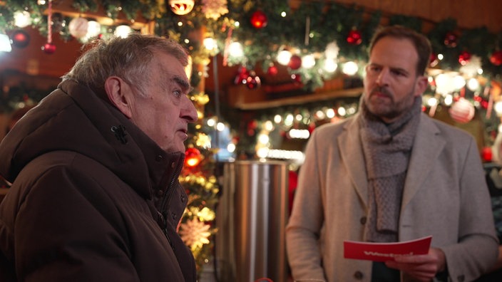 Herbert Reul und Henrik Hübschen auf dem Weihnachtsmarkt im Interview. Im Hintergrund ein Glühweinstand.