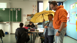 Jan-Niclas in seinem Klassenraum mit seinem Schulbegleiter. Jan-Niclas hält einen gelben Regenschirm hoch. Im Hintergrund sitzen anderen Kinder einem Schultisch.