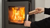 Eine Flamme lodert in einem Kaminofen, während eine Hand von rechts ein weiteres Stück Holz in den Ofen gibt,