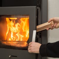 Eine Flamme lodert in einem Kaminofen, während eine Hand von rechts ein weiteres Stück Holz in den Ofen gibt,