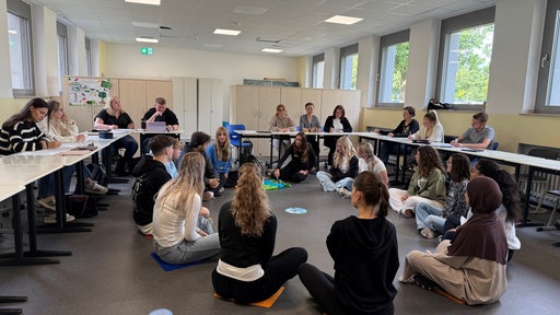 Junge Erzieher sitzen in einem Kreis und an Tischen in einem Klassenraum und reden