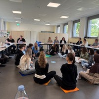 Junge Erzieher sitzen in einem Kreis und an Tischen in einem Klassenraum und reden