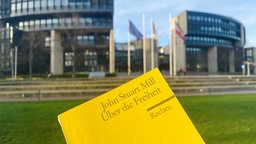 Das gelbe Reclam-Heft "Über die Freiheit" von John Stuart Mill im Vordergrund, im Hintergrund das Gebäude des Landtags in Düsseldorf