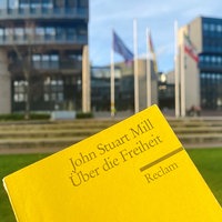 Das gelbe Reclam-Heft "Über die Freiheit" von John Stuart Mill im Vordergrund, im Hintergrund das Gebäude des Landtags in Düsseldorf