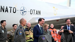 NRW- Ministerpräsident Hendrik Wüst und NATO-Generalsekretär Mark Rutte auf der Air Base in Geilenkirchen. Sie stehen vor einem Flugzeug von Leuten umgeben in Uniformen und halten Uniformen in der Hand. Beide lachen.