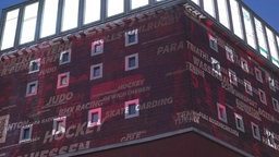 An eine rotbraune Hausfassade wird Schrift projiziert, sie zählt Sportarten auf.