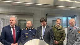 Minister Optendrenk in der Gereon-Kaserne. Er steht mit vier weiteren Männern in einer Küche und sie schauen in einen leeren Suppentopf.