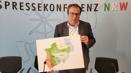 Ein Mann im Anzug hält eine NRW-Karte hoch mit einer farbigen Einteilung
