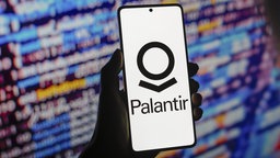 Name und Logo des Softwareunternehmens Palantir auf einem Smartphone - im Hintergrund verschwommen ein Bildschirm mit bunten Elementen wie Zahlen und Buchstaben.