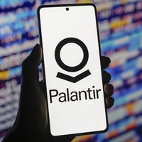 Name und Logo des Softwareunternehmens Palantir auf einem Smartphone - im Hintergrund verschwommen ein Bildschirm mit bunten Elementen wie Zahlen und Buchstaben.