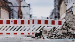 Eine Straße mit Häusern rechts und Links. Rcehts im Vordergrund ein kleiner Schutthaufen und die Straße ist abgetrennt von einem rot-weißen Bauzaun.