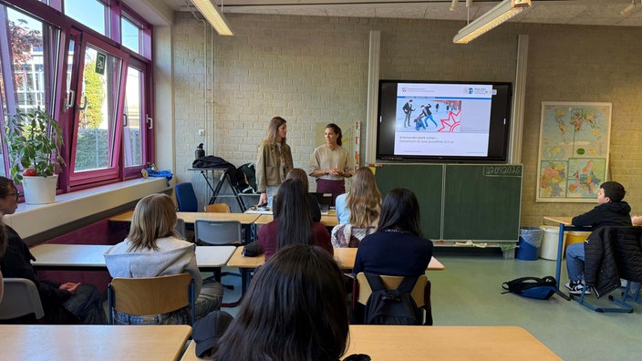 Zwei Polizistinnen in Zivil stehen vor einer Schulklasse