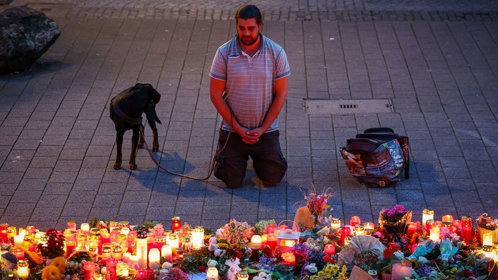 Ein Mann kniet trauernd vor einer großen Menge Kerzen und Blumen. Neben ihm steht sein Hund.