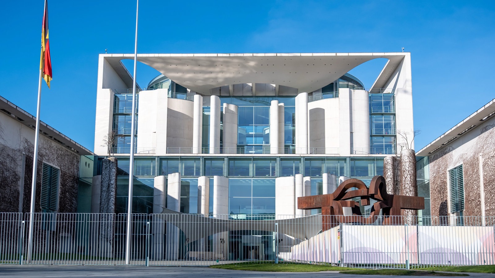 Das Kanzleramt in Berlin, links hängt eine Deutschlandfahne, der Himmel ist strahlend blau.