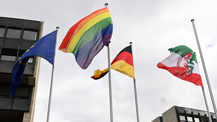Vor dem Landtag in Düsseldorf weht eine Regenbogenflagge neben einer Deutschlandflagge, einer Europaflagge und einer Flagge mit dem NRW-Landeswappen.