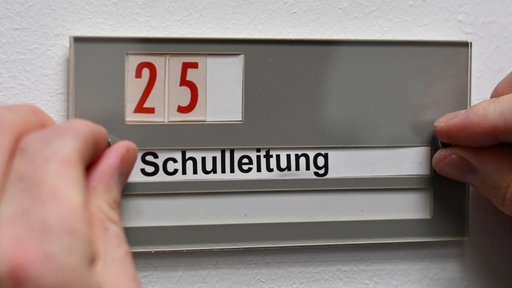 Zwei Hände befästigen ein Schild an der Wand mit der Aufschrift: Schulleitung und der Nummer 25 