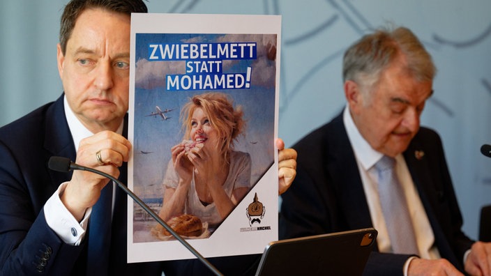 Jürgen Kayser (l), Leiter des NRW-Verfassungsschutzes, und NRW-Innenminister Herbert Reul (CDU) bei der Vorstellung des NRW Verfassungsschutzberichts 2025. Kayser hält dabei ein Plakat mit einem KI Bild aus dem Rechten Spektrum.
