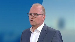 EIn Mann mit schütterem Haar, runder dunkler Brille und Anzug vor einem blauen Hintergrund