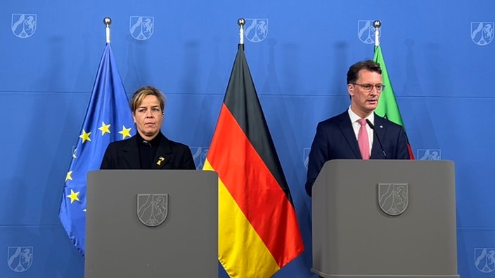 Mona Neubaur und Hendrik Wüst stehen nebeneinander vor Redepulten und geben eine Pressekonferenz