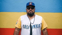 Benny Bajo mit einem Sporttrikot, Sonnebrille und Baseballcap vor einer blau-gelb-rot gestreiften Wand