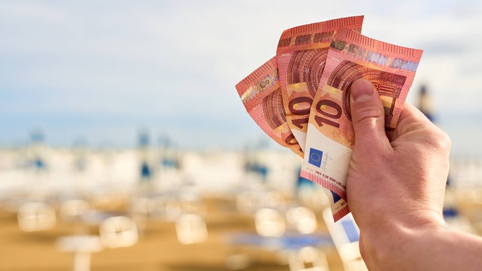 Ruka drži nekoliko novčanica, u pozadini prazna plaža