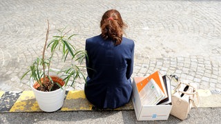 Eine Frau sitz auf dem Gehweg. Neben ihr ihre Tasche und einen Karton voller Arbitsmappen