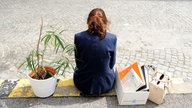 Eine Frau sitz auf dem Gehweg. Neben ihr ihre Tasche und einen Karton voller Arbitsmappen
