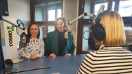 Dwie rozmówczynie przy mikrofonie w studiu radiowym, a plecami siedzi prowadząca