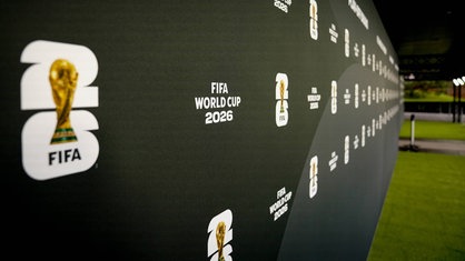 Symbolbild: dunkle Begrenzungs-Wand in einem Stadion, darauf Logos der FIFA mit dem WM-Pokal in einer stilisierten 26, im Hintergrund rechts Rasenfläche des Spielfelds
