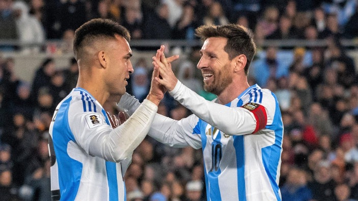 Länderspielszene: Die argentinischen Nationalspieler Lionel Messi (r., mit Kapitänsbinde) und Lautaro Martínez (l.) kommen freudig zueinander und verschränken je eine ihrer gehobenen Hände, im Hintergrund unscharf Zuschauerränge
