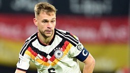 Länderspielszene: Joshua Kimmich läuft im DFB-Trikot mit Kapitänsbinde bei einem Länderspiel engagiert über den Platz (Nahaufnahme), im Hintergrund unscharf eine große Deutschlandflagge in den Zuschauerrängen (November 2025, dreiviertel-Portrait)