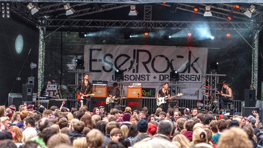 Eine Band steht vor Publikum auf einer Bühne, im Hintergrund das Banner: "EselRock, umsonst + draußen"
