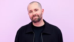 DJ David Puentez mit dunkler Kleidung, steht vor einem rosa Hintergrund