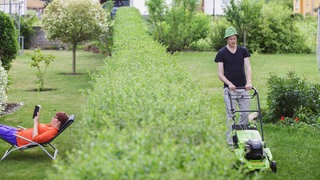 Ein Mann mäht seinen Rasen, während seine Nachbarin hinter der Hecke in einem Liegestuhl ein Buch liest.