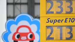 Ein Schild mit einem stilisierten Auto mit Mund und Augen ist hinter den Preisen für Benzin (E10) und Diesel zu sehen. 