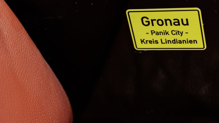 Ein Aufkleber mit der Aufschrift "Gronau - Panik City - Kreis Lindianien" auf der Couch im Hotelzimmer aus Hamburg in der Sonderausstellung "Udo L. wohnt im Hotel - Exklusive Einblicke in meine Panikzentrale" im Rock'n'Popmuseum in Gronau.