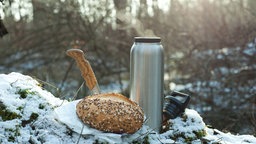 Ein Brötchen, ein Messer und eine Thermosflasche mit einem heißen Getränk stehen für ein Picknick in einem verschneiten Wald