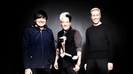 Die Ärzte stehen vor einem dunklen Hintergund mit einem Koffer und lächeln in die Kamera