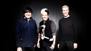 Die Ärzte stehen vor einem dunklen Hintergund mit einem Koffer und lächeln in die Kamera
