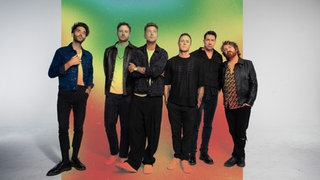 Die Bandmitglieder stehen vor einer bunten Wand.