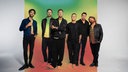 Die Bandmitglieder stehen vor einer bunten Wand.