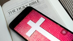 Eine gedruckte Bibel auf der ein Smartphone liegt, auf dessen Bildschirm ein großes Kreuz prangt.