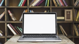 Auf einem Schreibtisch aus Holz steht ein aufeklappter Laptop. Der Bildschirm ist leer. Im Hintergrund ist ein Regal mit Büchern.