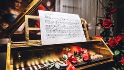 Ein goldenes Tasteninstrument steht geöffnet da. Auf den Tasten liegen rote Rosen. Ein handgeschriebenes Notenblatt lehnt oben am Instrument. Im Hintergrund sorgen weitere Rosen und warmes Licht für eine festliche, romantische Stimmung.