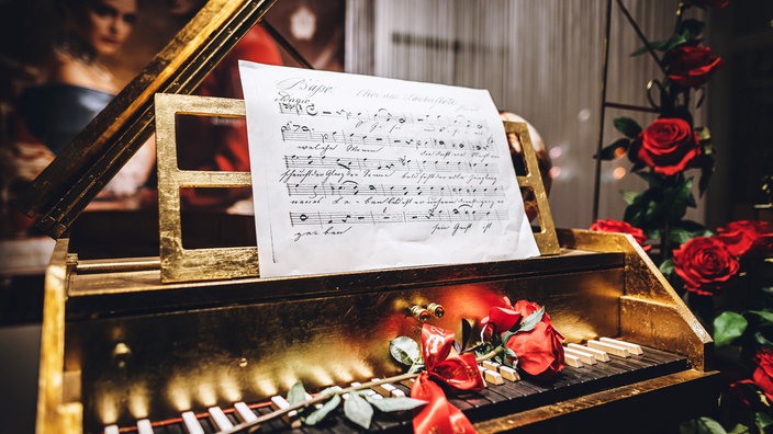 Ein goldenes Tasteninstrument steht geöffnet da. Auf den Tasten liegen rote Rosen. Ein handgeschriebenes Notenblatt lehnt oben am Instrument. Im Hintergrund sorgen weitere Rosen und warmes Licht für eine festliche, romantische Stimmung.